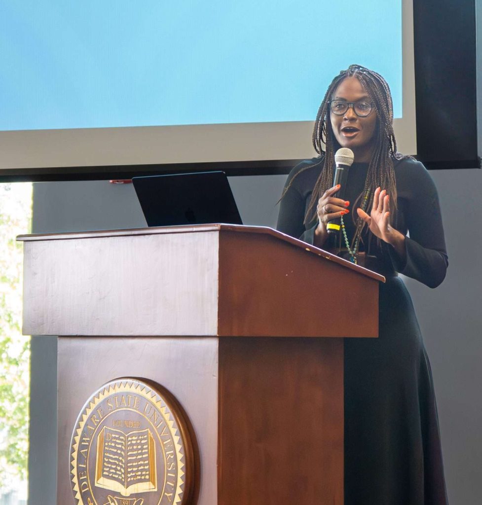 A woman in a black dress speaks at a podium with a university seal, presenting in front of a large screen displaying partial text. A laptop is open on the podium.