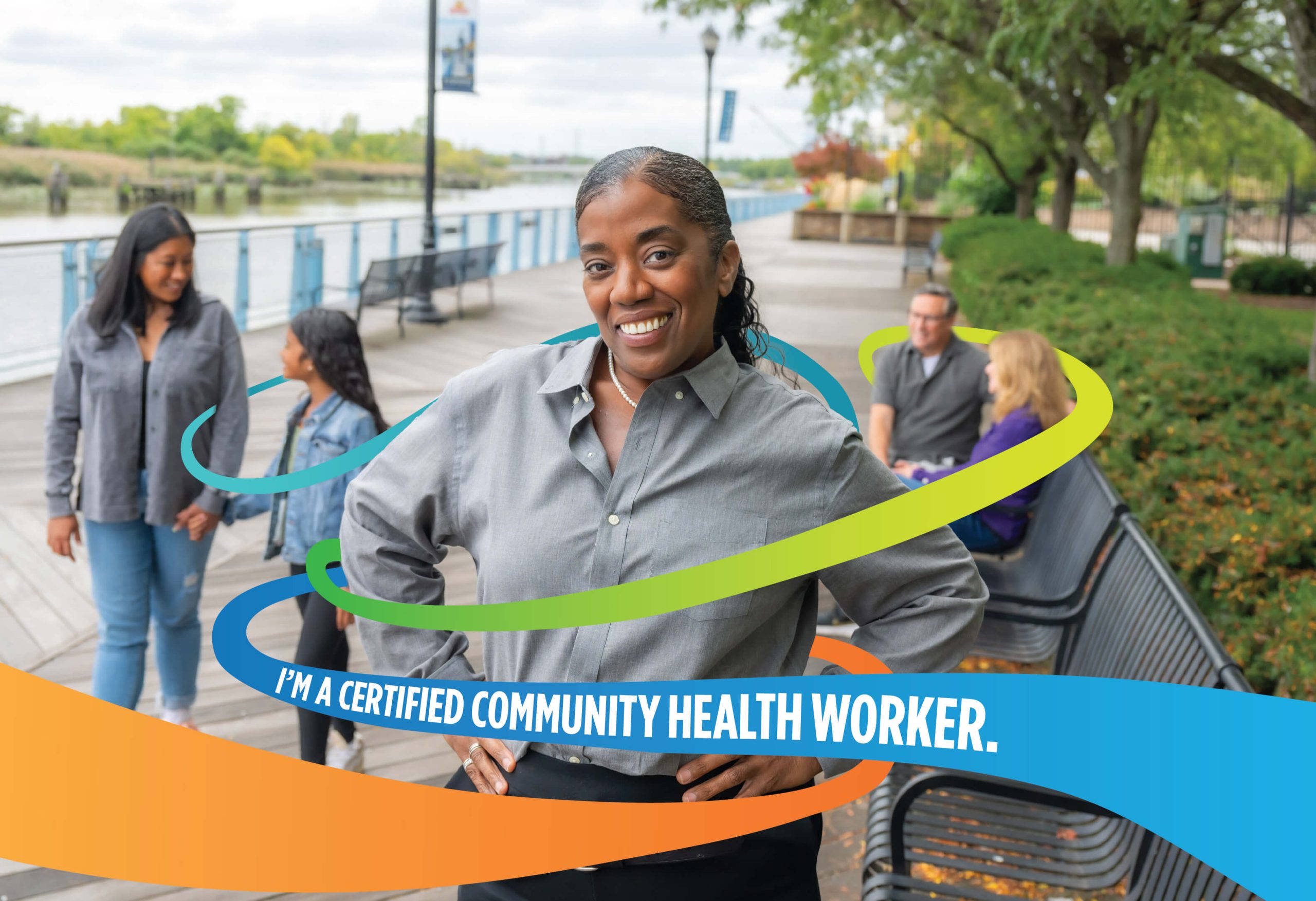 A smiling woman stands outside with her hands on her hips, surrounded by three other people. Colorful swooshes and text read, I’m a certified community health worker. Trees and a river are in the background.